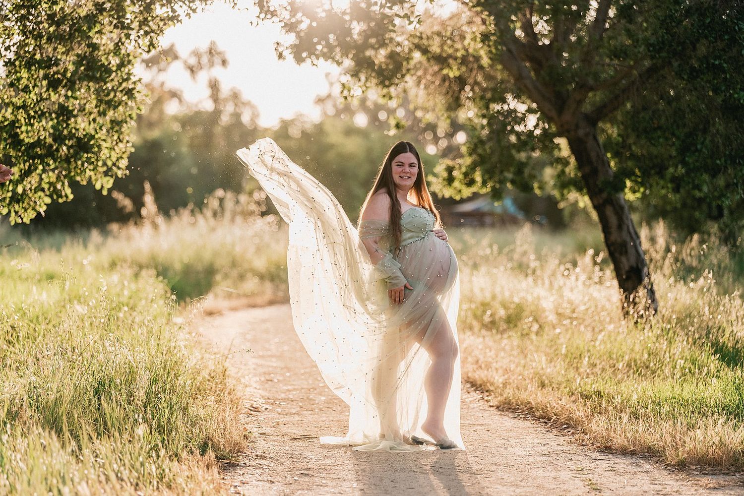 Smiling pregnant woman in flowing white lace maternity dress posing on a sunny outdoor path with trees.