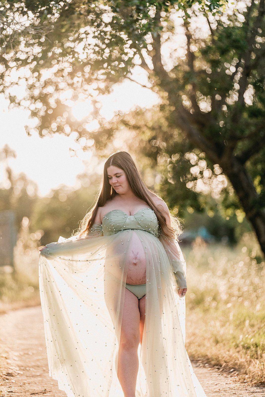Pregnant woman in sage green lingerie and sheer cape poses outdoors in golden sunlight among trees.
