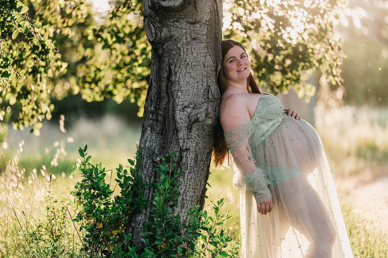 Pregnant woman in sheer green gown leaning against tree in sunlit meadow during golden hour maternity photo session.