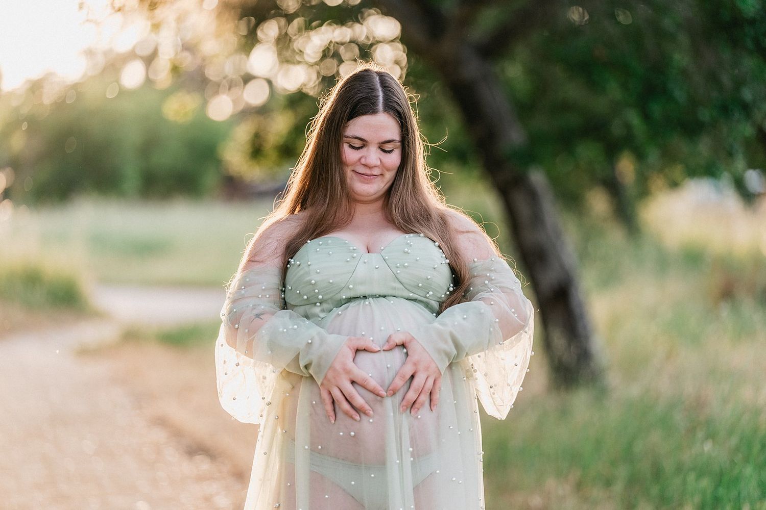 Pregnant woman in sage green maternity gown making heart shape over belly outdoors at golden hour.