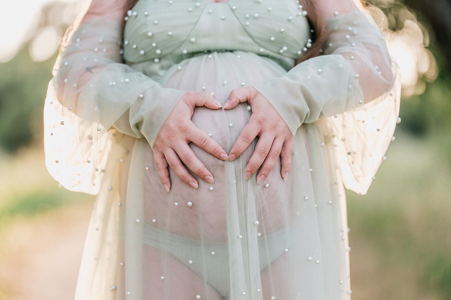 Pregnant woman in pearl-embellished tulle gown forming heart shape with hands on baby bump outdoors.