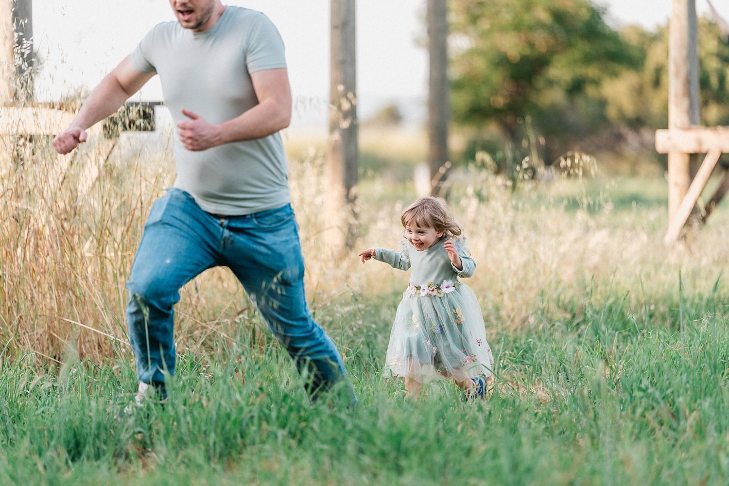 A father and toddler daughter playing and running together in a green meadow field during golden hour.
