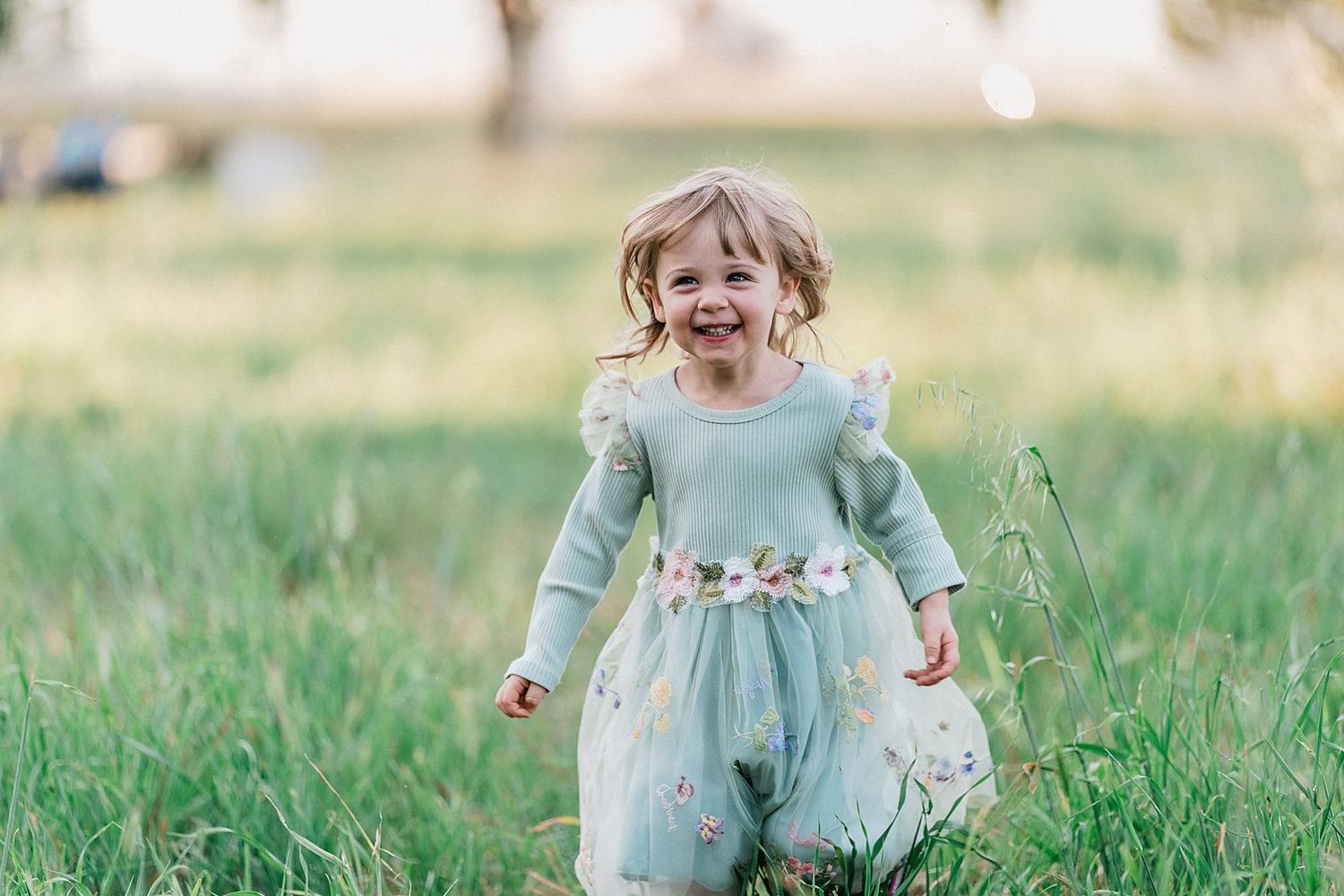 A joyful toddler girl in a mint green floral dress runs through a lush green field, laughing with delight.