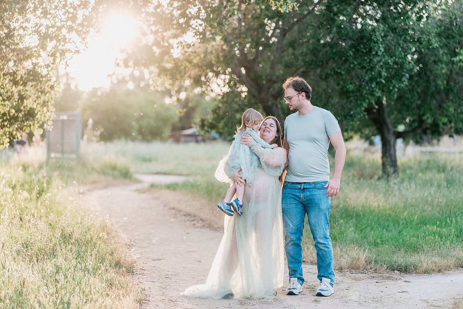 Pregnant woman in sheer gown with husband and child on sunny dirt path surrounded by green trees.