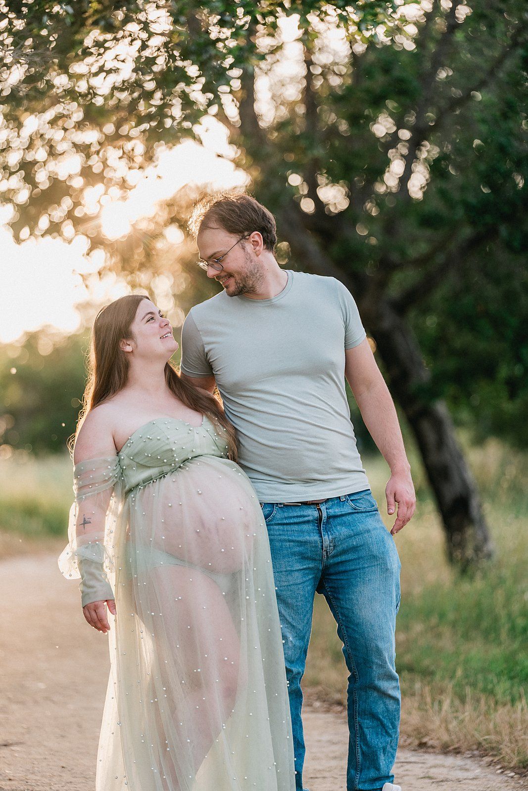 Expecting couple sharing a smile while walking on a sunlit path with lush trees in background.