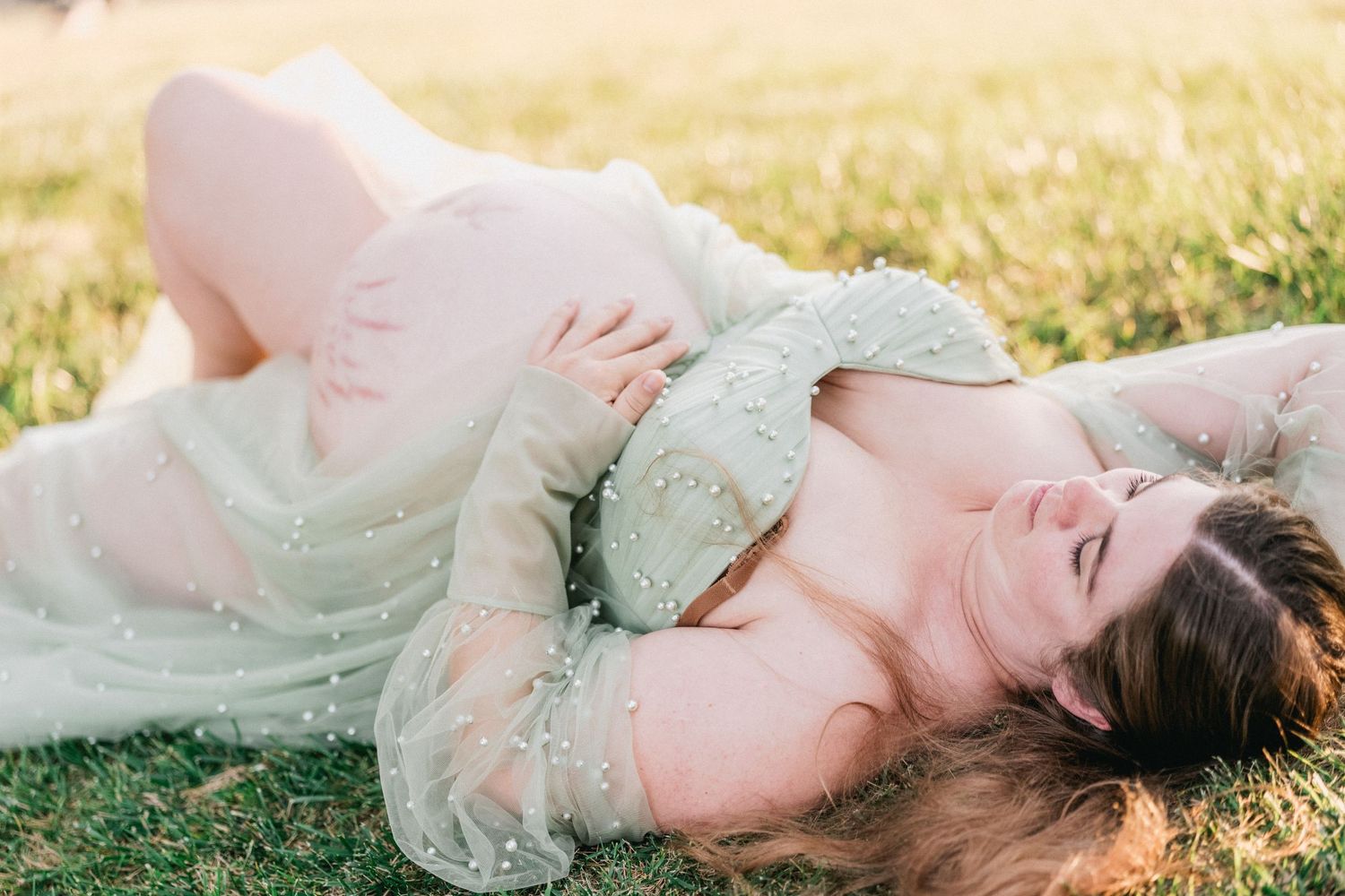 Pregnant woman in elegant beaded green gown lying on grass, cradling baby bump during outdoor maternity photo session.