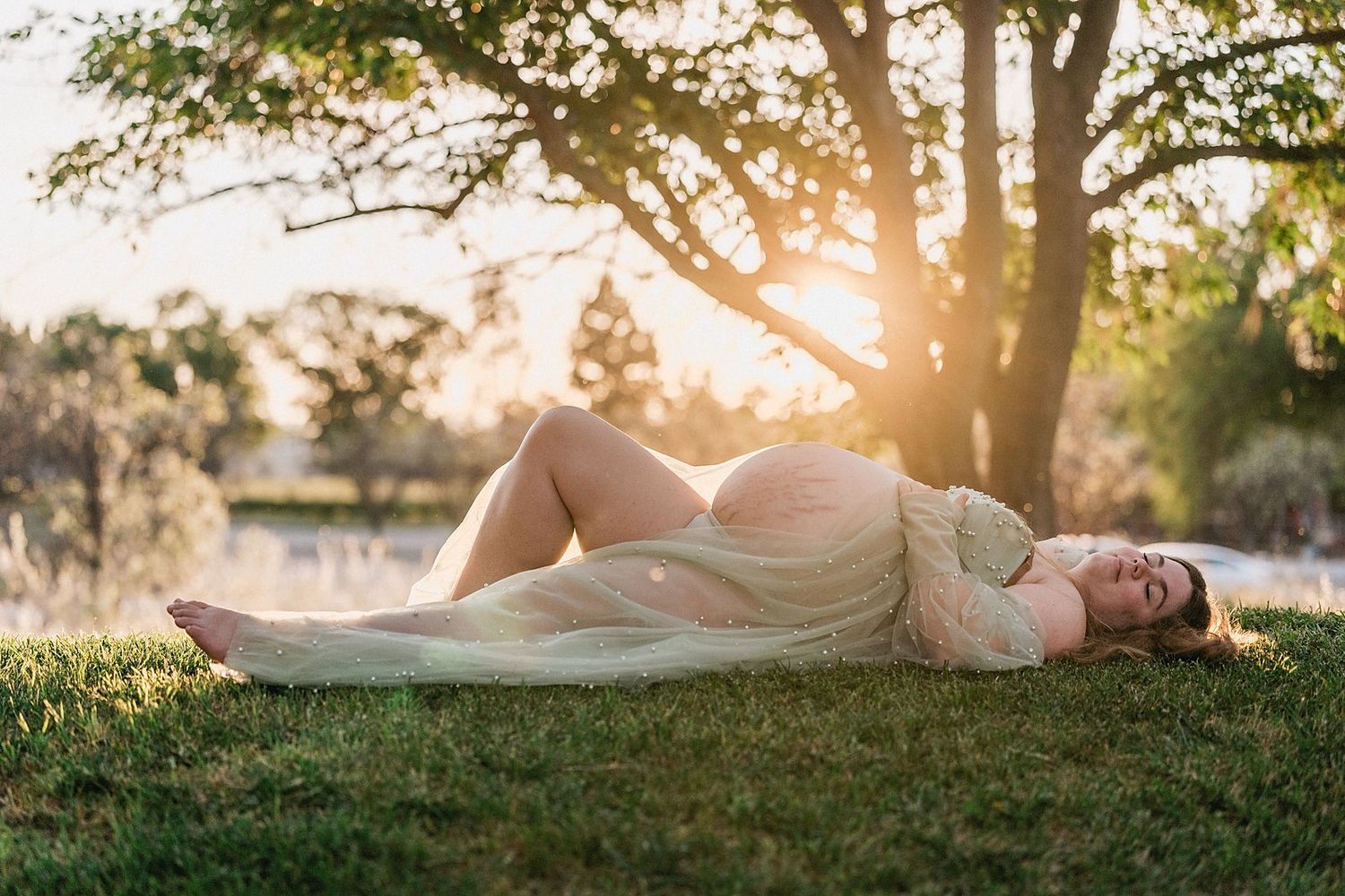 A pregnant woman in a flowing dress lying on grass under a tree in warm sunset light.