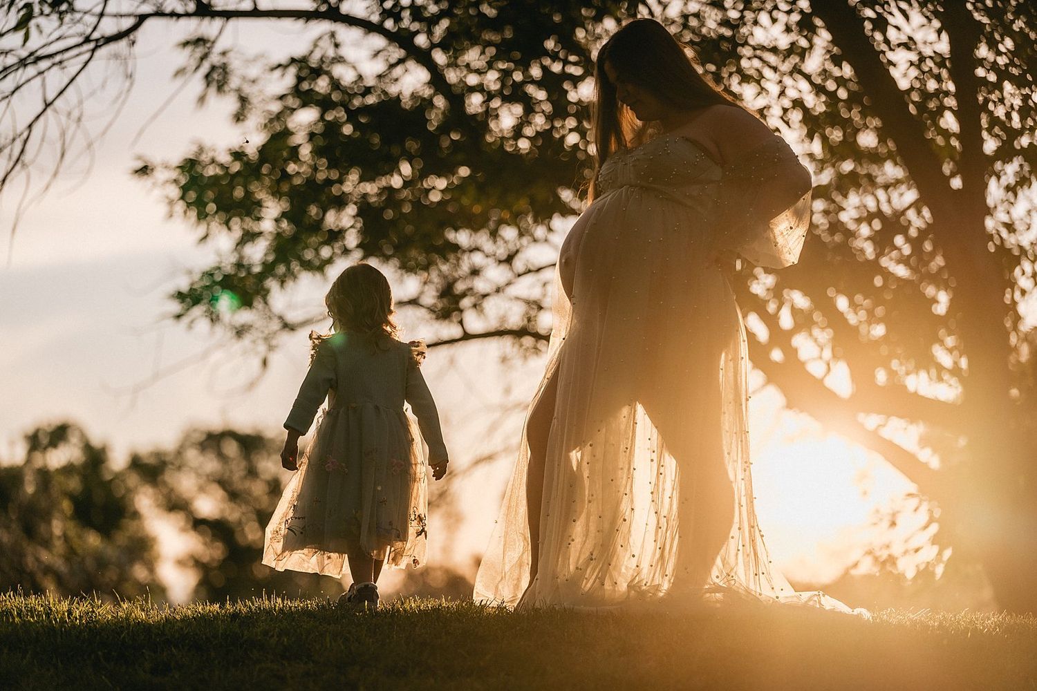 Silhouette of pregnant woman and young girl walking under trees at golden sunset.