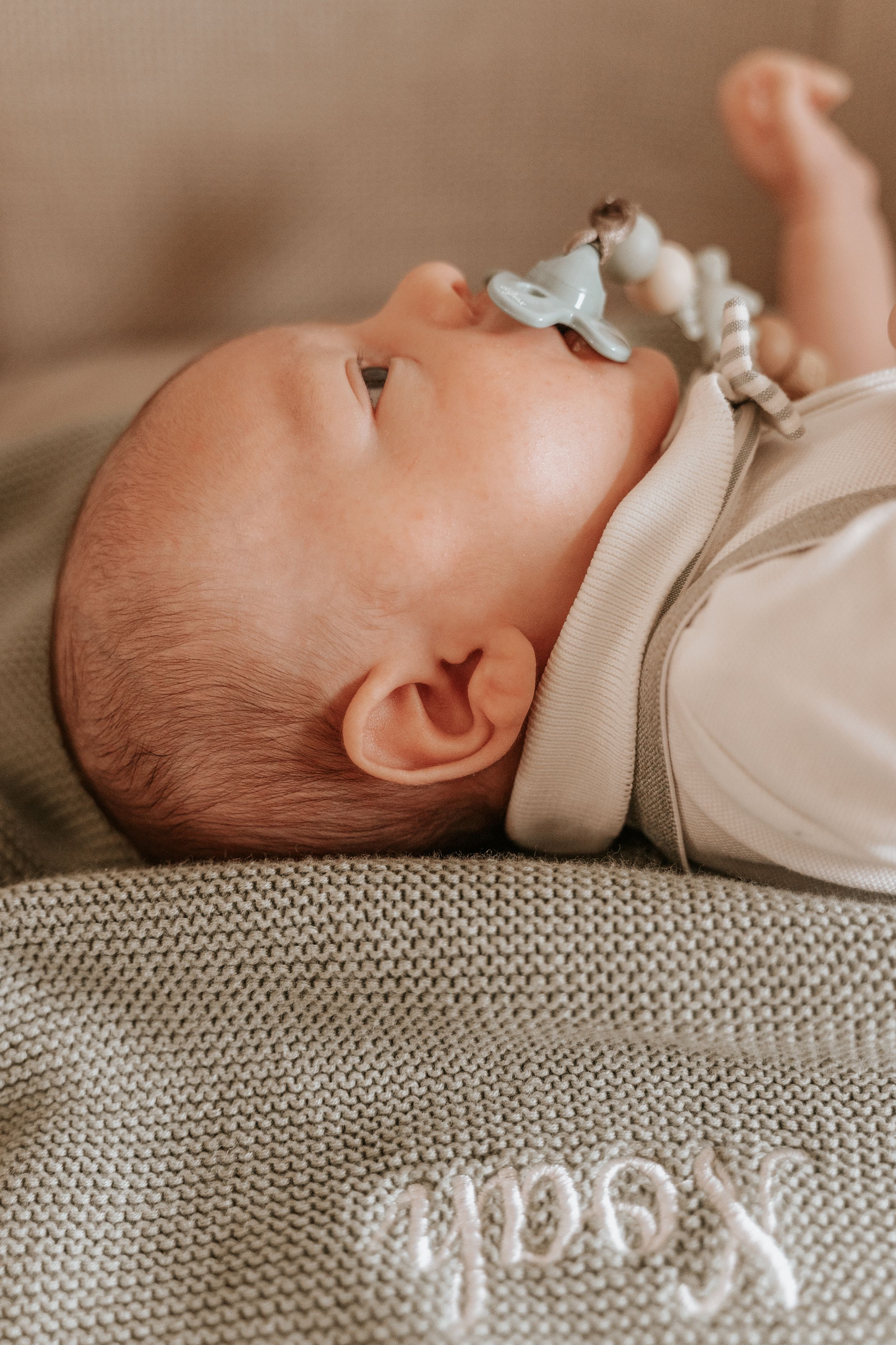 Newborn baby lying on a knitted blanket embroidered with the name Noah, holding a pacifier clip.