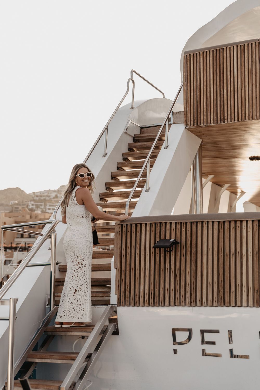 A person in a white lace dress stands on the deck of a luxury yacht against a wooden slatted wall.