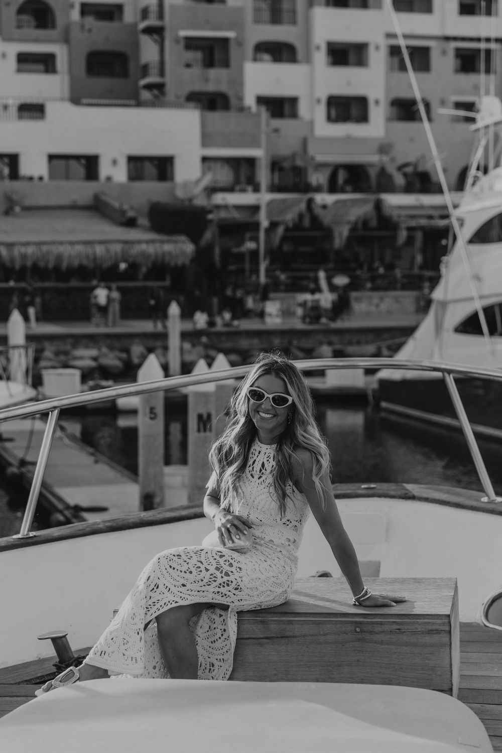 Woman in white lace dress and sunglasses poses on luxury yacht dock in black and white photograph.