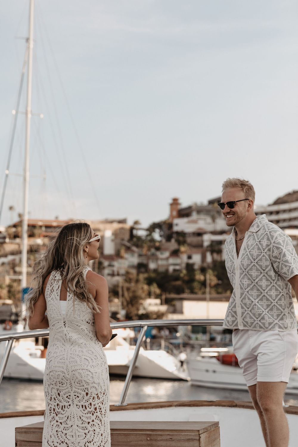 Couple in matching white lace outfits pose together on a marina dock with boats and coastal buildings in background.