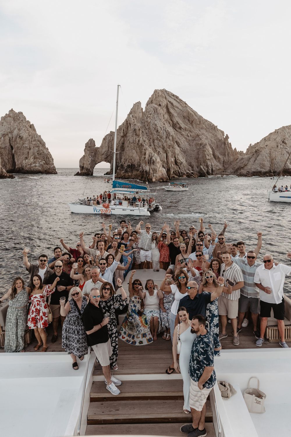Large group celebrating on a boat deck with El Arco rock formation visible in the background at Cabo San Lucas.