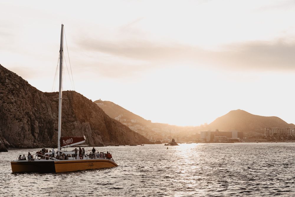 Panoramic sunset view of mountains and water with a sailboat silhouetted against the golden sky.