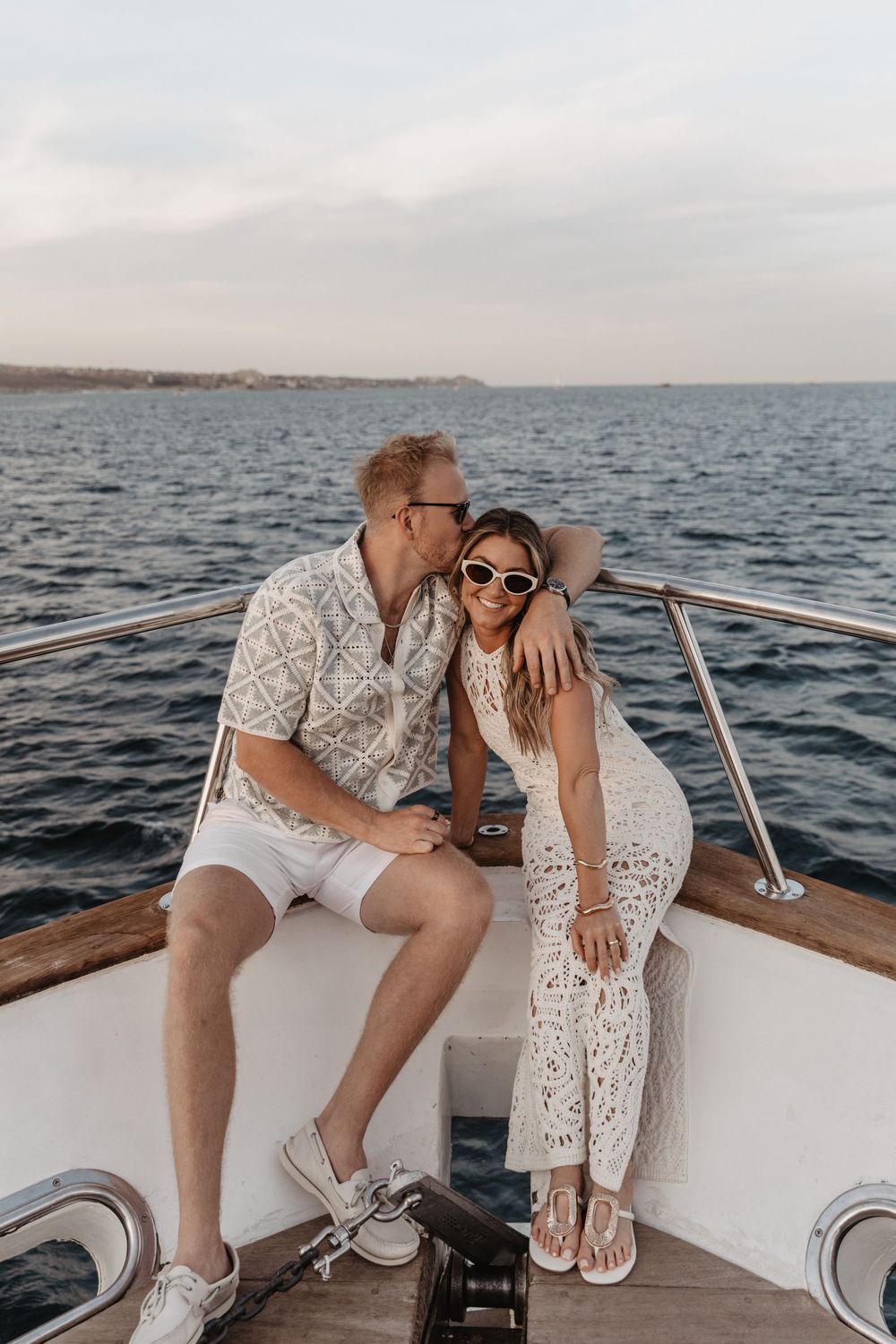 Couple in white lace outfits sharing romantic moments aboard a boat at sunset on calm waters.