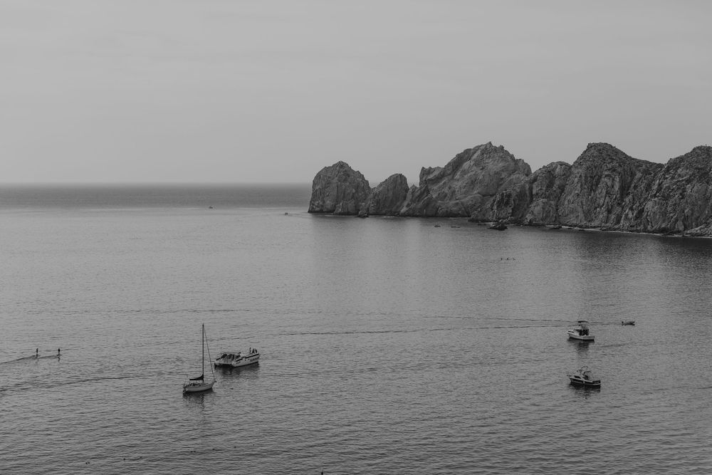 Sailboats anchored in calm waters near rocky coastal cliffs in black and white photography.