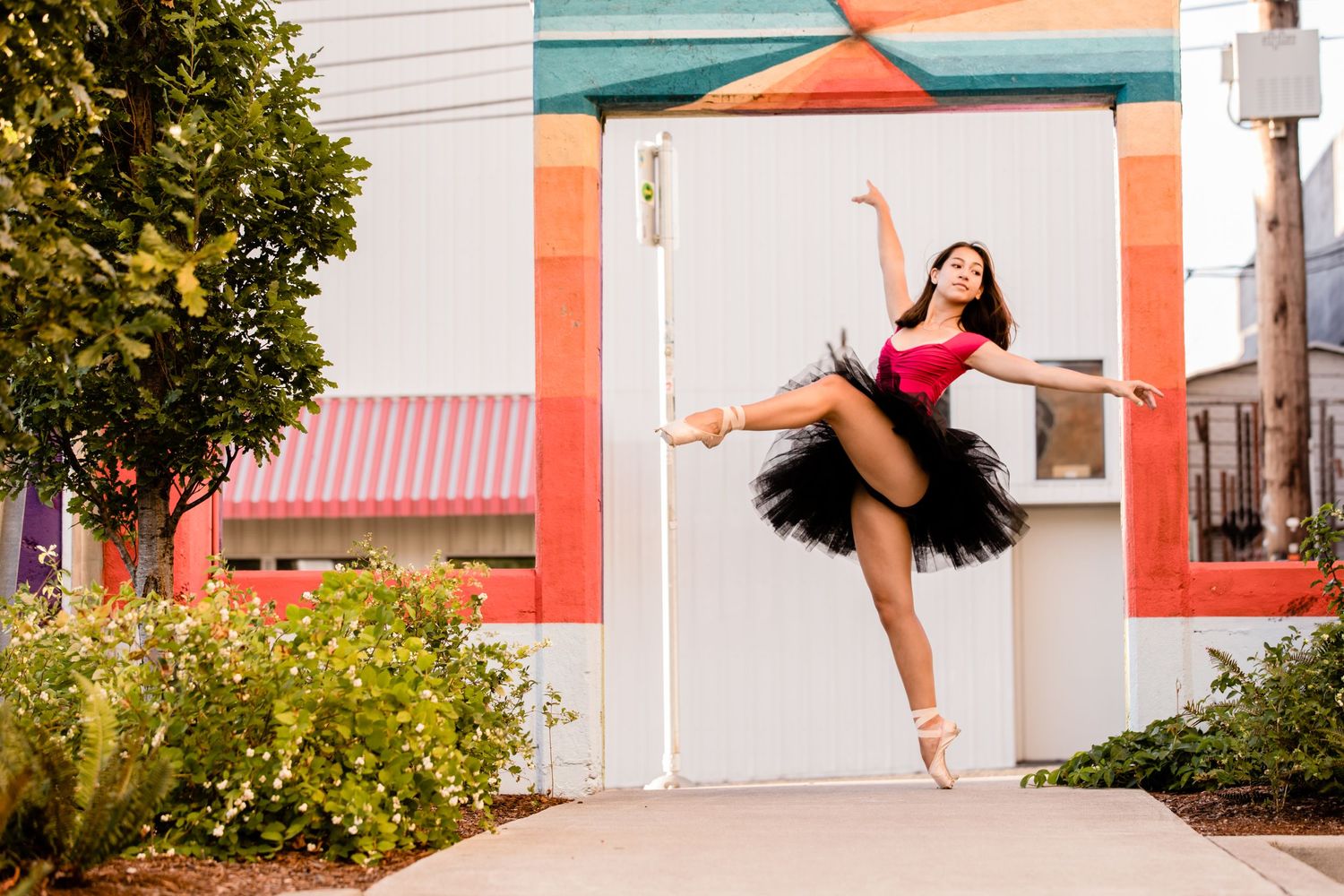 A dancer in a red top and black tutu performs an elegant arabesque pose against a pastel-colored wall outdoors.