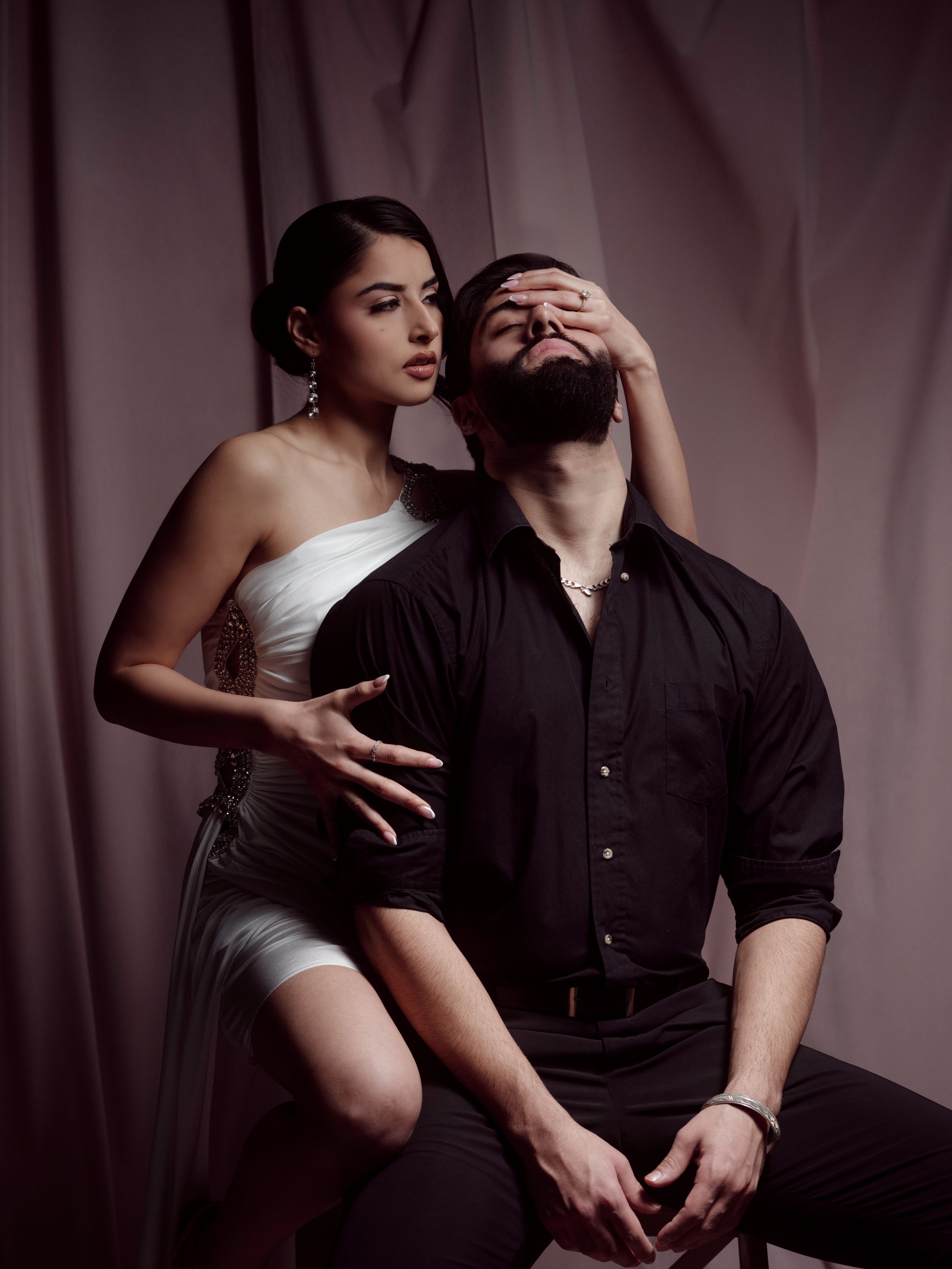Artistic portrait in moody lighting shows couple in intimate pose against pink backdrop, wearing white and black attire.
