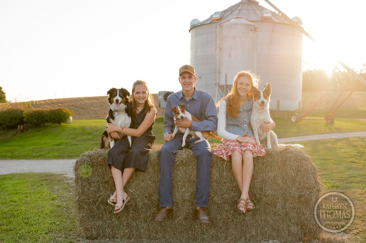 Group photo with dogs sitting on hay bales in front of grain silo at sunset.