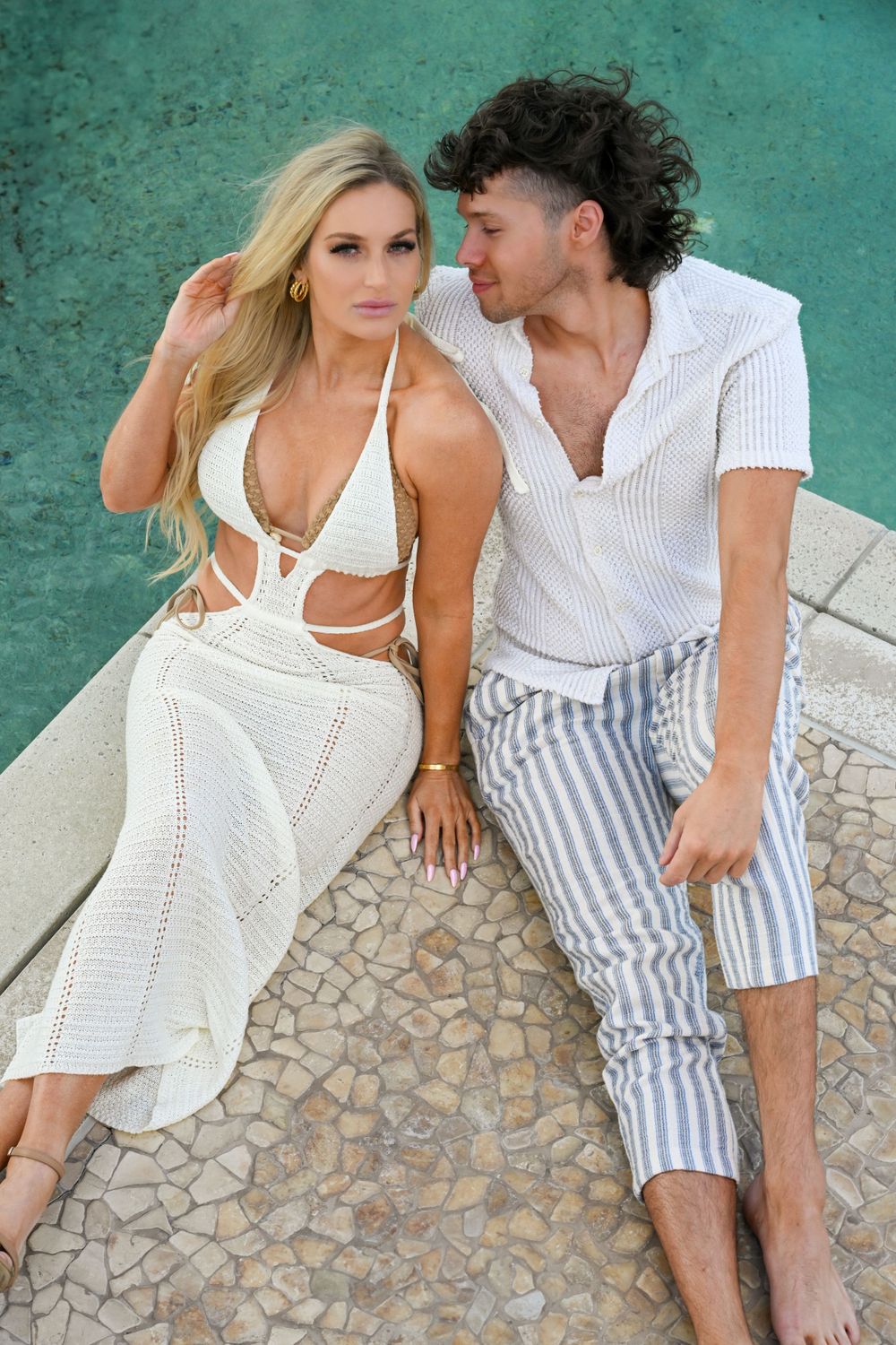 Couple in white and striped summer outfits pose against turquoise wall backdrop.