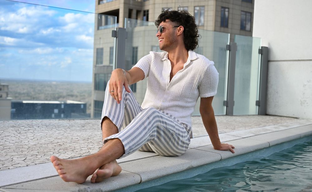 A person in a white shirt and striped pants relaxes by the edge of a rooftop pool with a cityscape view.