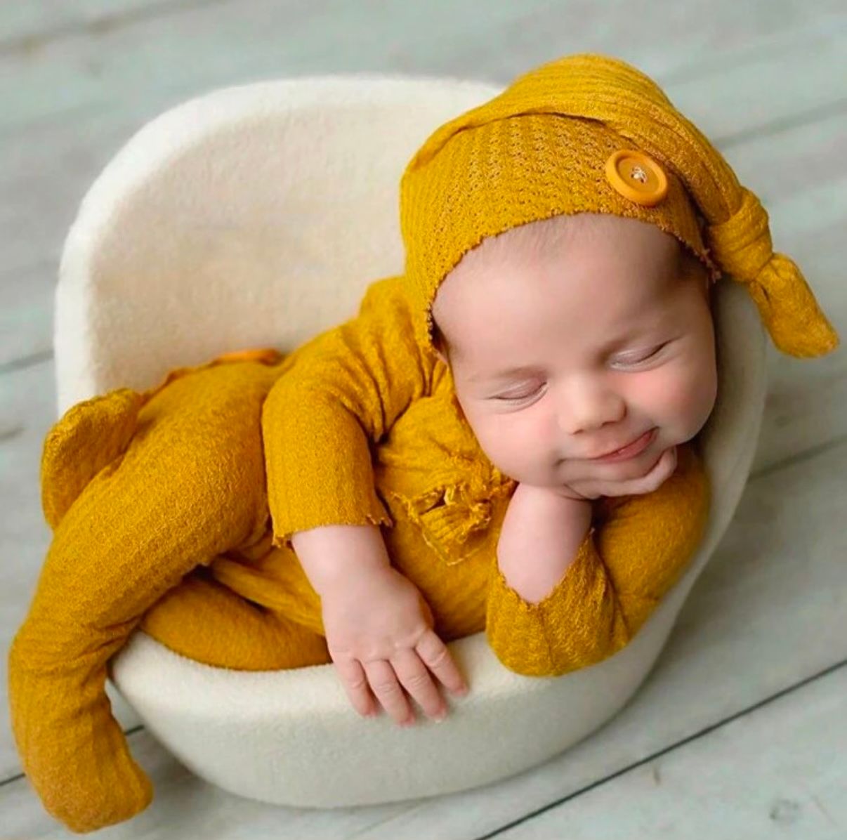 Sleeping newborn in mustard yellow knit outfit and matching hat on white cushion.