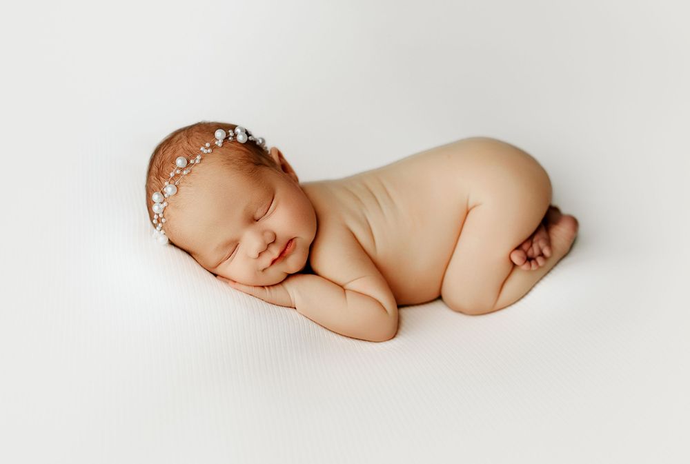 A sleeping newborn baby wearing a delicate pearl headband curled up on a soft white background during a photography session in Portsmouth OH