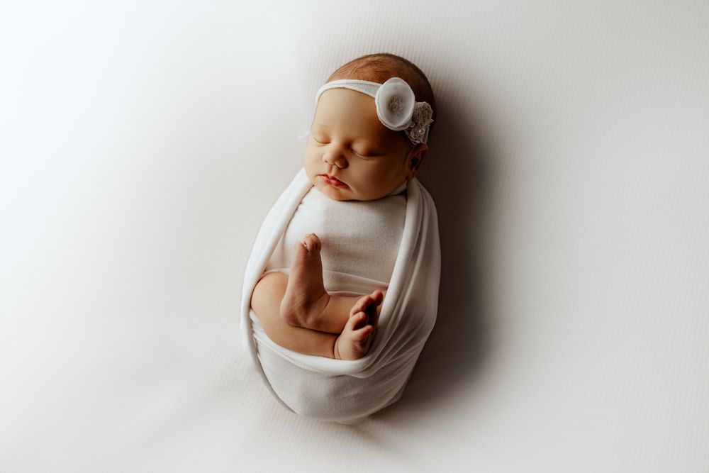 A sleeping infant swaddled in white cloth with a white headband lies on a plain white background.