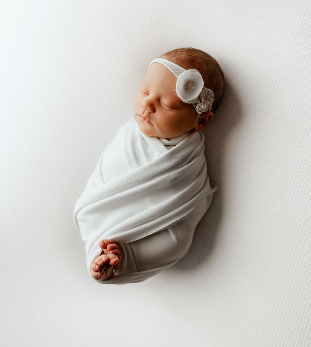 Sleeping newborn baby swaddled in white fabric and wearing a light headband lies peacefully against white background.