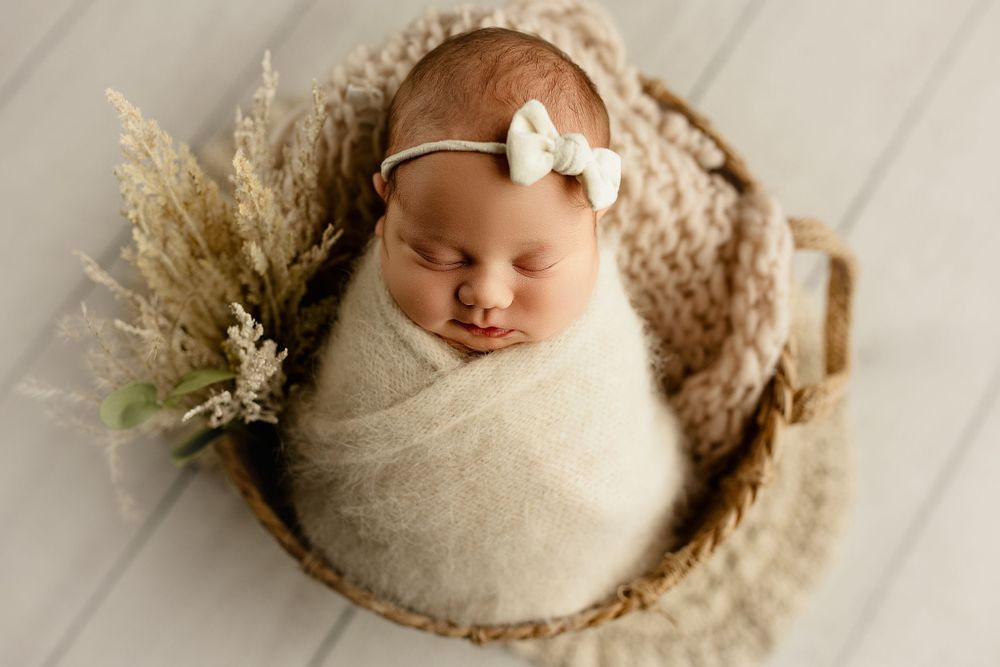 Newborn baby peacefully sleeping in cozy basket with dried flowers and cream wrap, wearing white bow headband.