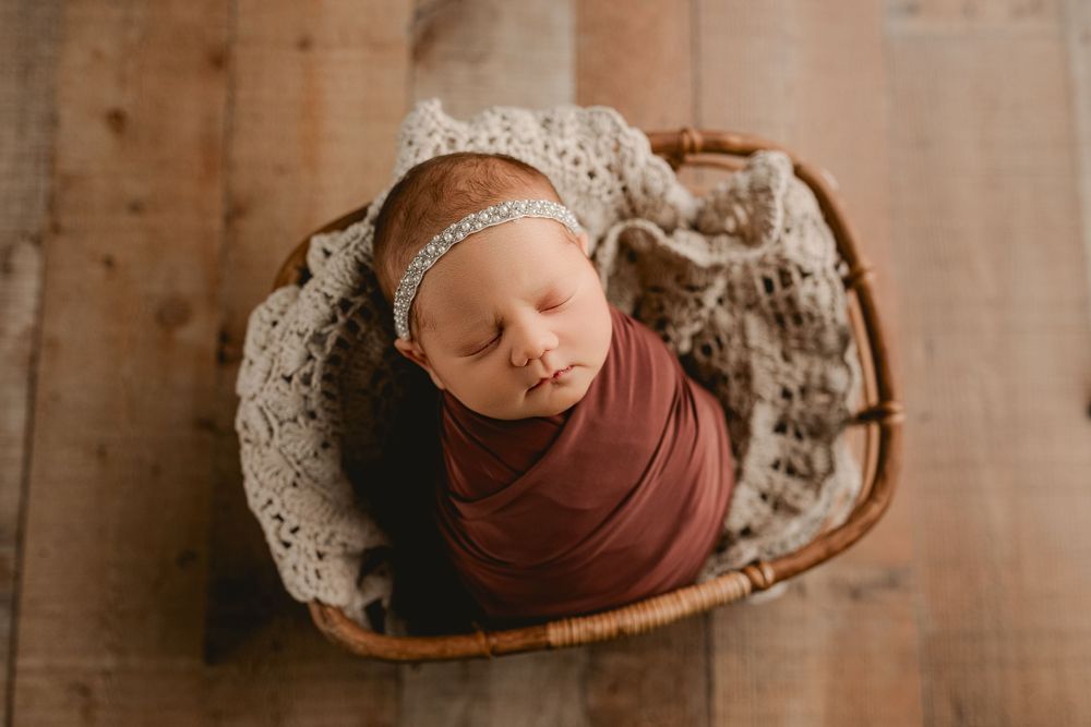 Newborn baby swaddled in brown fabric sleeping peacefully in a rustic basket with lace trim and headband.