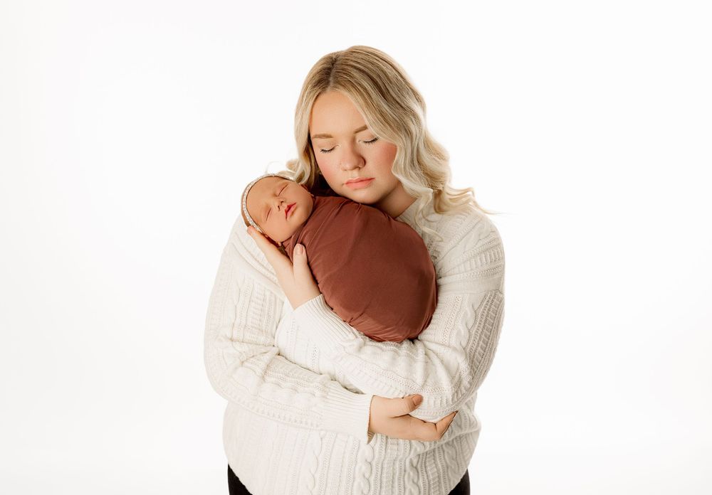 Mother in cream sweater cuddles newborn wrapped in brown fabric against white backdrop.