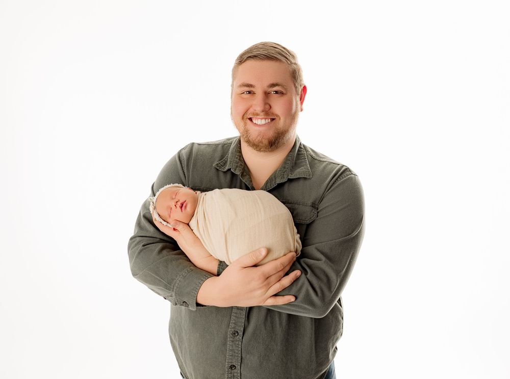 Parent holding sleeping newborn wrapped in cream fabric against white backdrop.