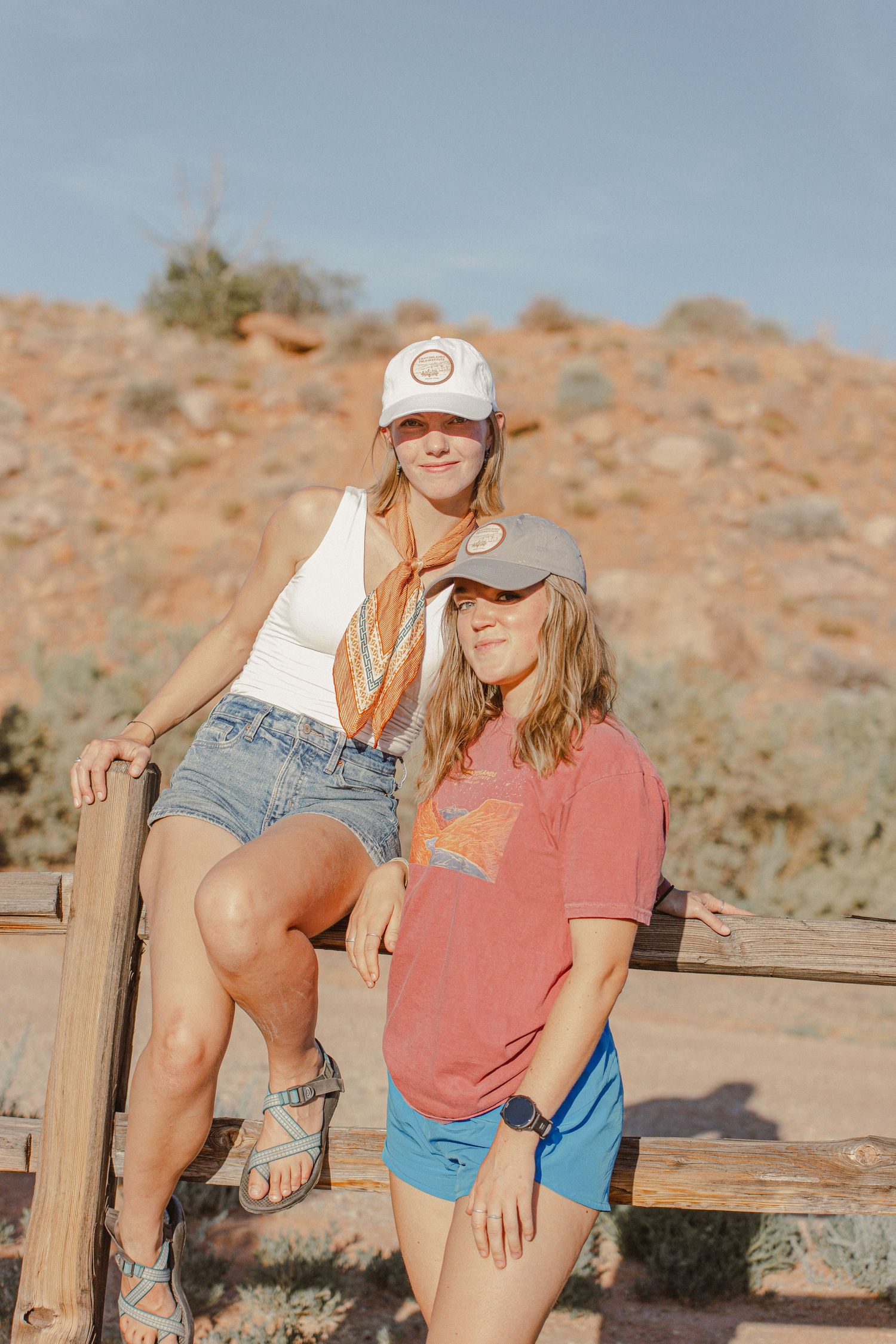 Two people in casual summer attire pose on wooden railing in desert landscape.