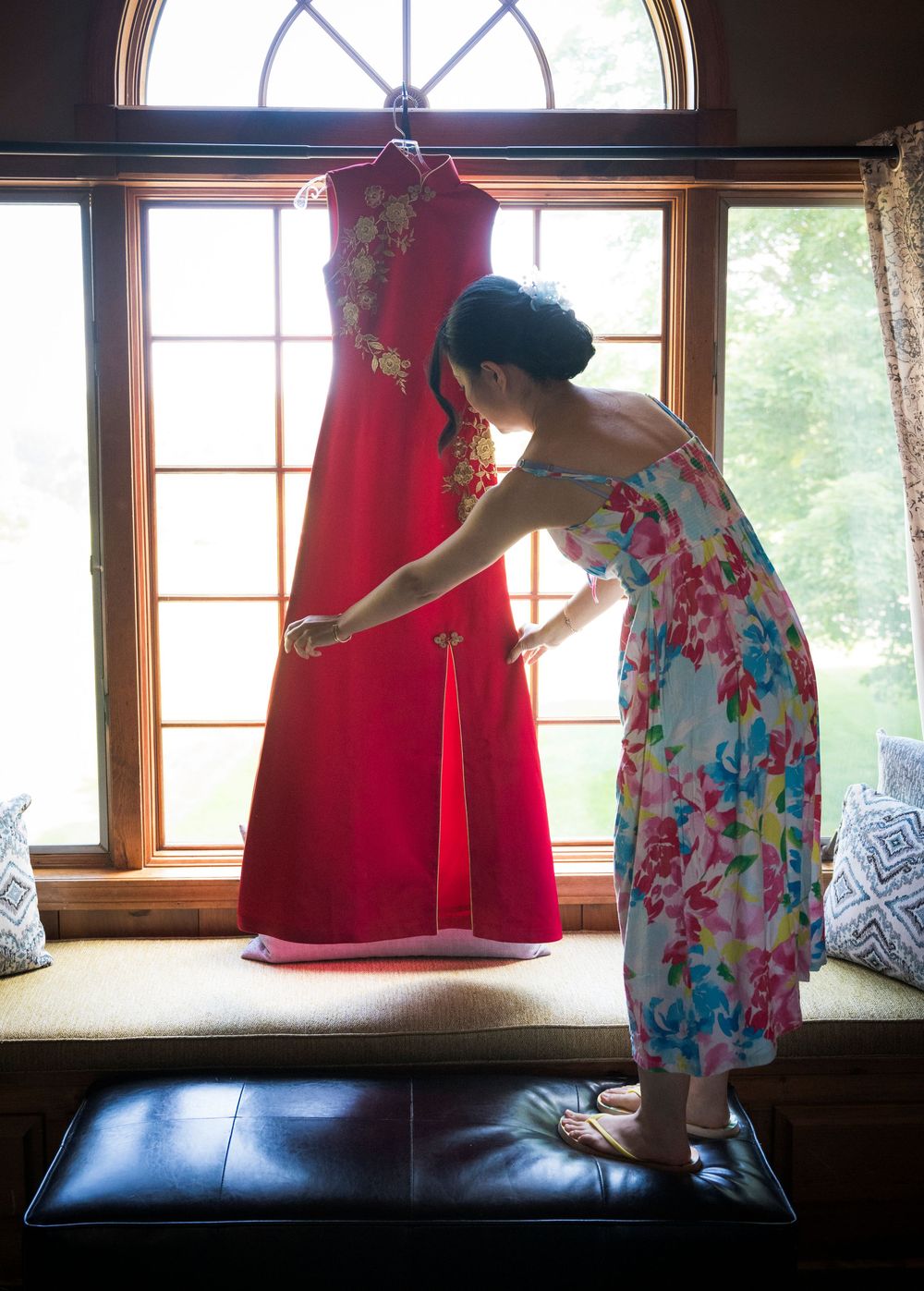 A floral dress-clad figure adjusts a hanging red formal gown near sunlit windows.