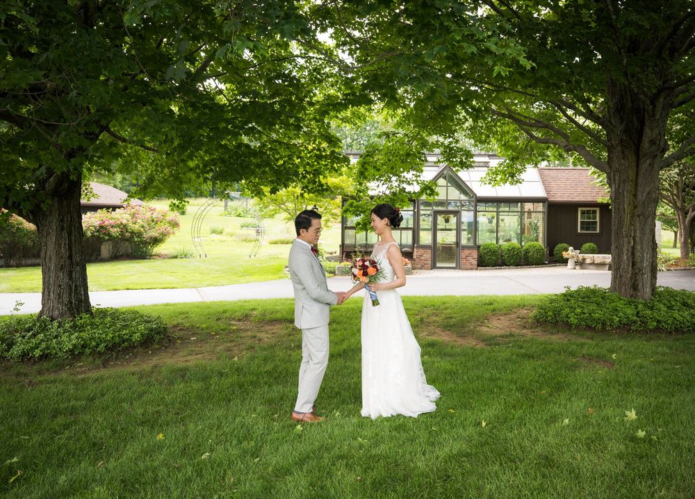A wedding couple shares a romantic moment under large shady trees on a lush green lawn near a country house.