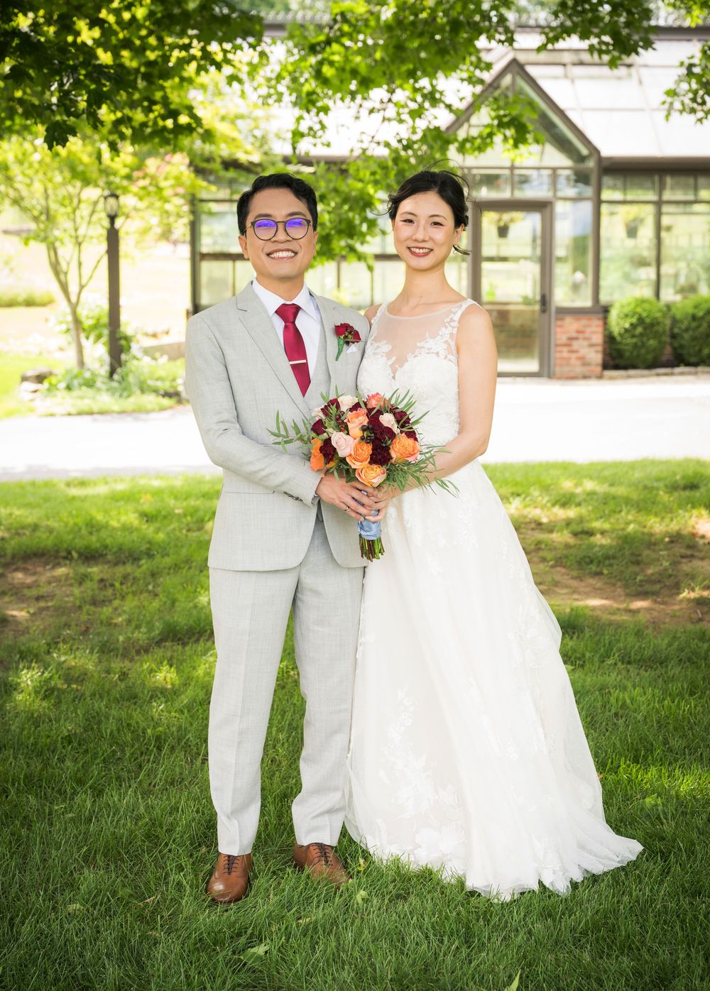 A couple poses outdoors in wedding attire with a greenhouse backdrop.