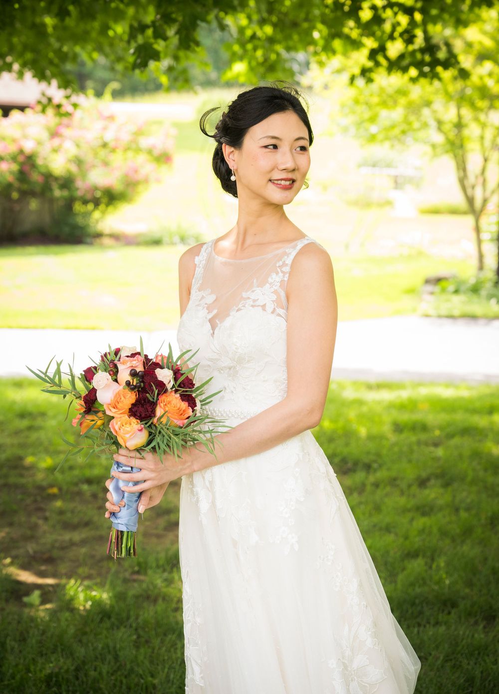 Bride in elegant lace wedding dress holds vibrant autumn-colored bouquet in sunny garden setting.