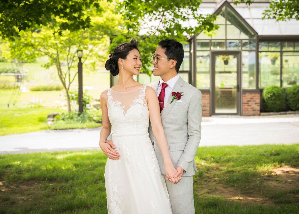 Newlyweds share a tender moment while holding hands in front of a glass greenhouse surrounded by greenery.