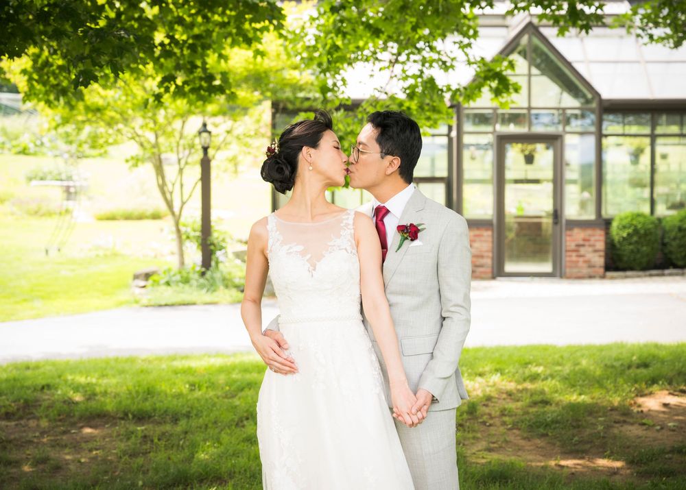 Couple in wedding attire standing together in front of a greenhouse on a sunny summer day.