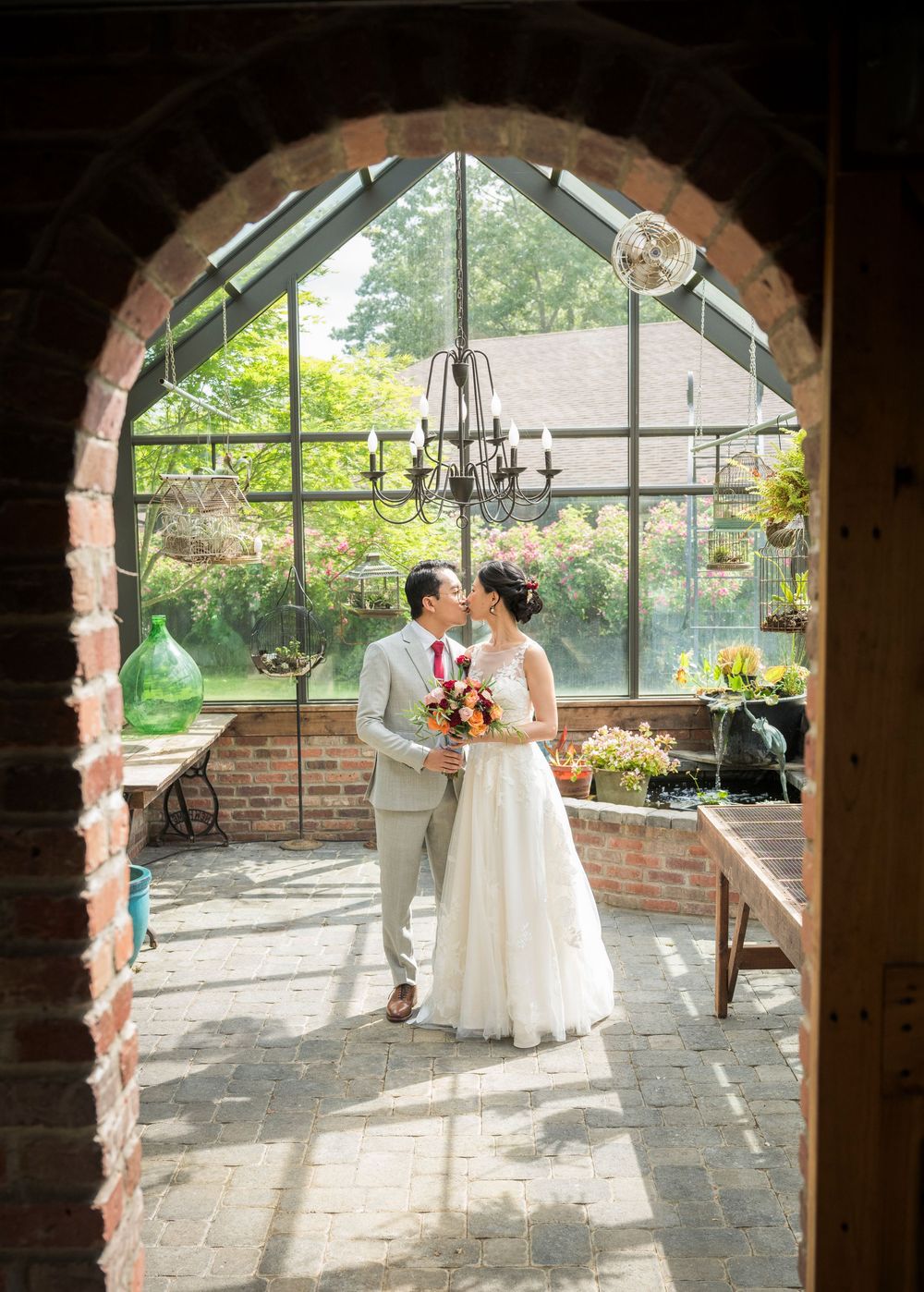 A sunlit greenhouse wedding ceremony space with brick archway, vintage chandelier and rustic decor.