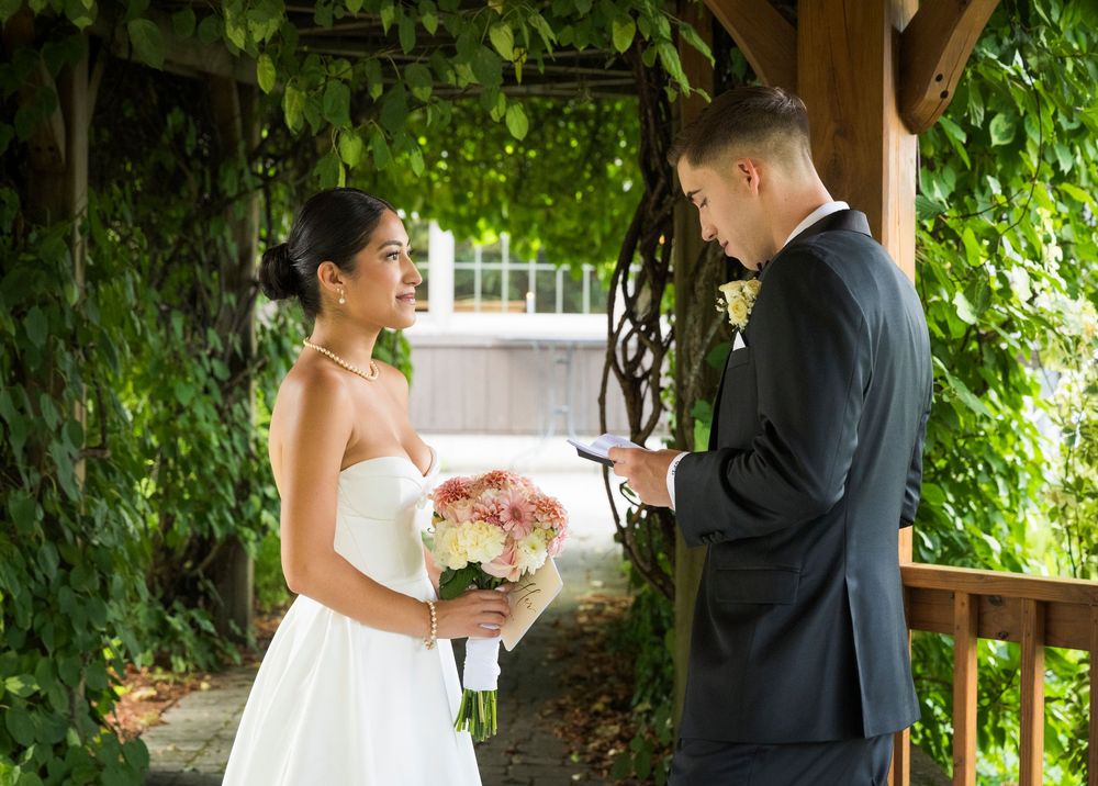 Wedding couple poses beneath wooden gazebo surrounded by lush greenery and flowers.