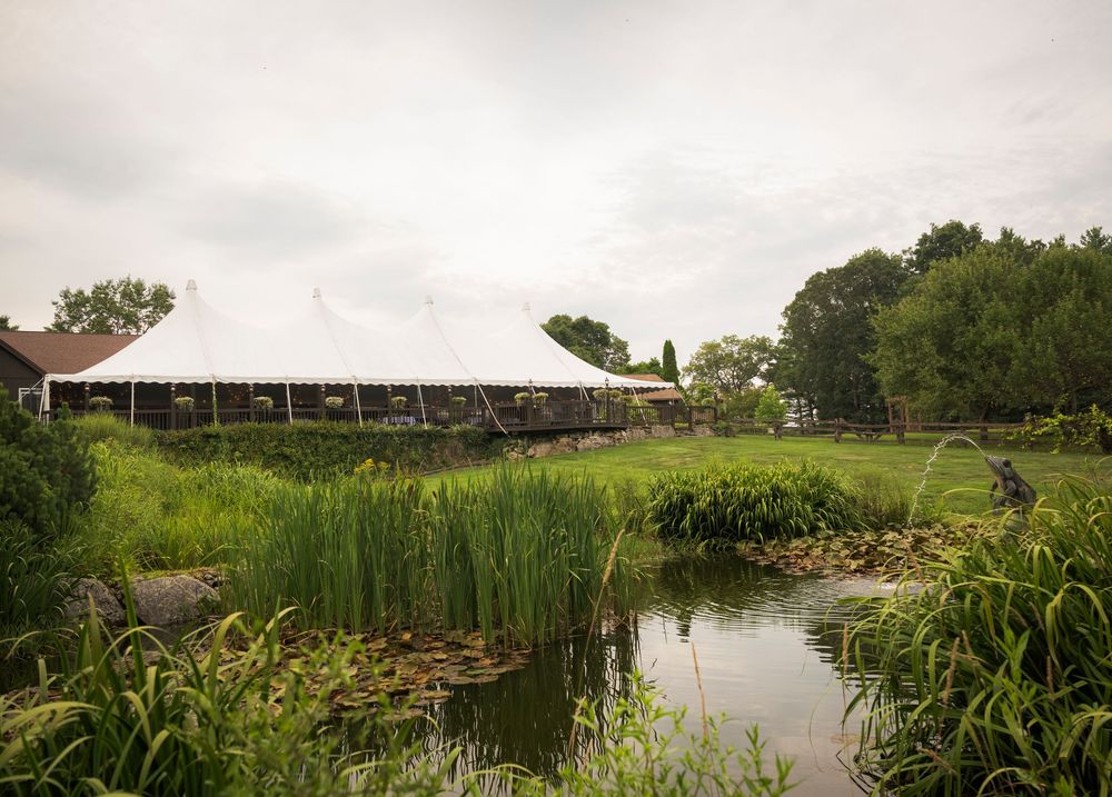 Scenic wedding venue landscape showing a serene pond with white tents and natural foliage in the background.