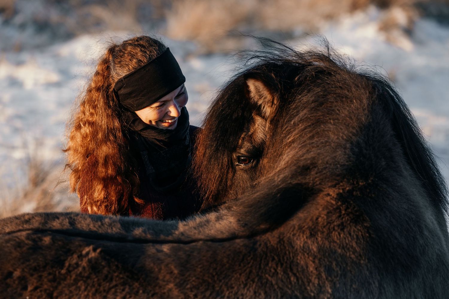 A person with curly red hair wearing a winter hat bonds with a dark-colored horse in warm sunset lighting.
