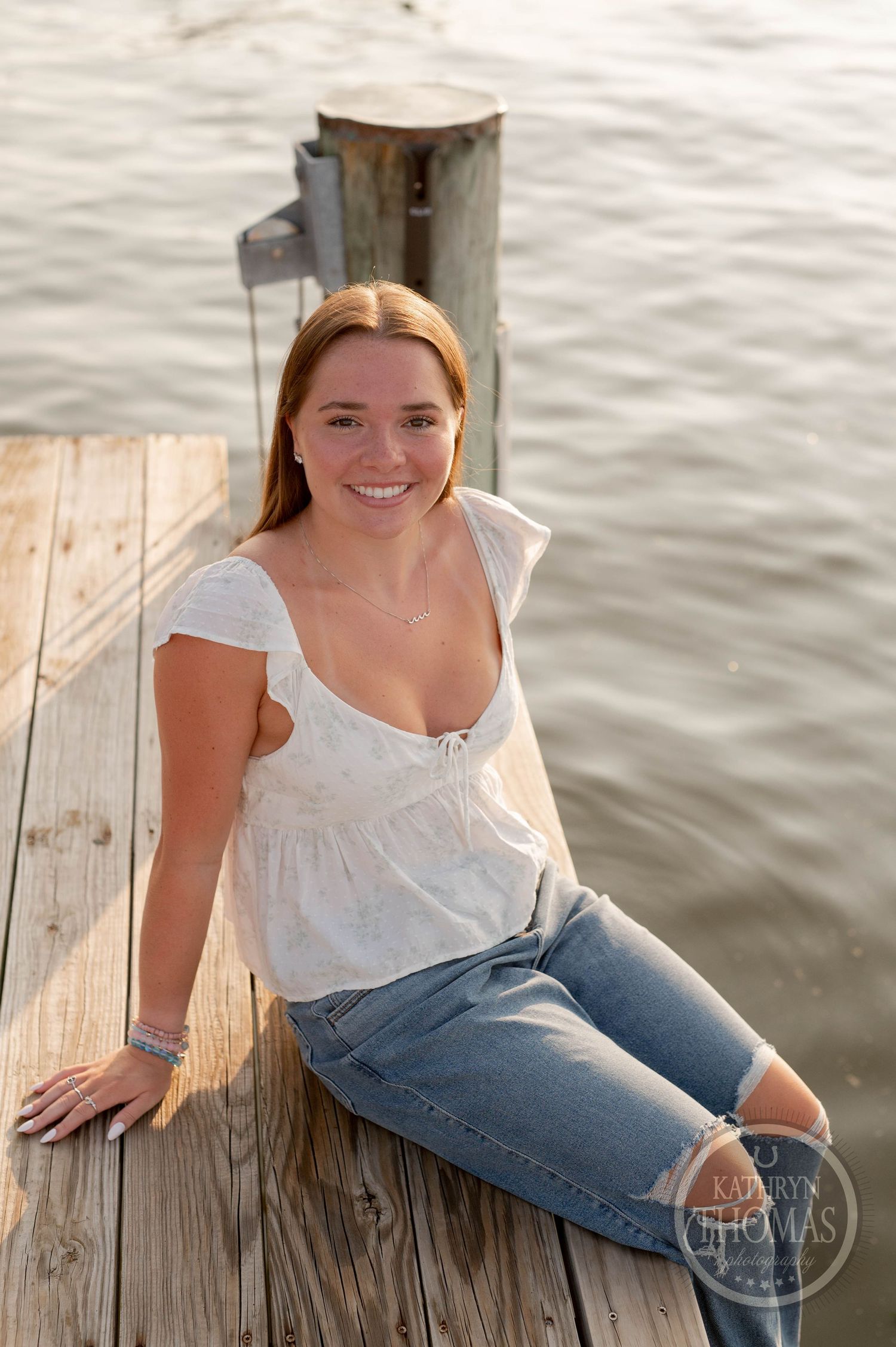 Person in white top and ripped jeans sitting on dock by water at sunset.