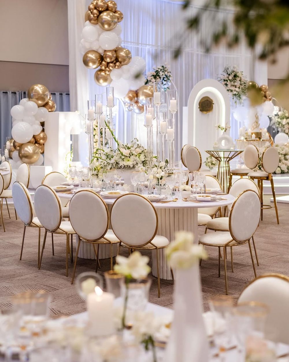 Elegant wedding reception setup with white and gold decor, round tables, plush chairs and balloon arrangements.