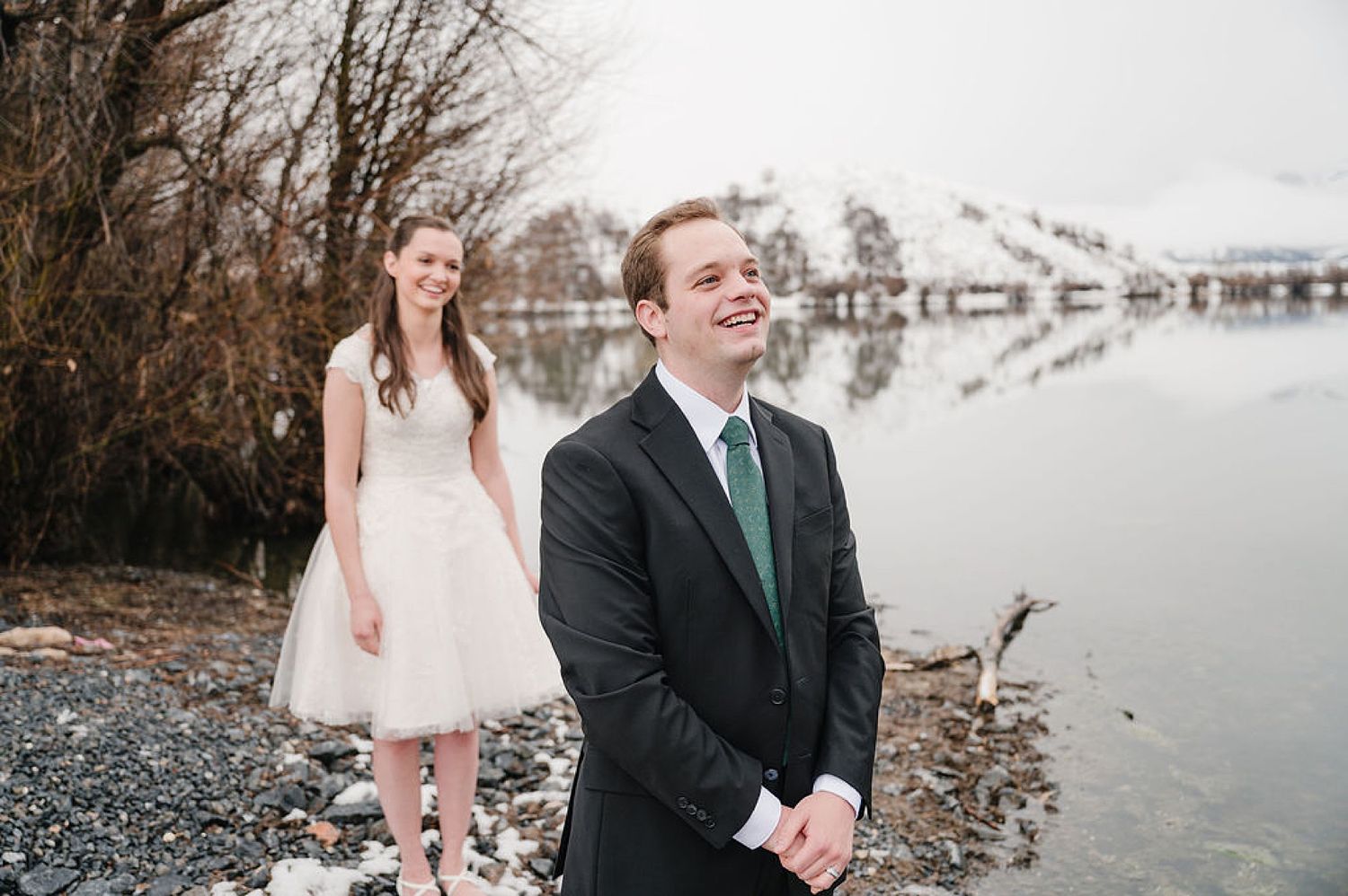A couple in wedding attire shares a joyful moment on a rocky lakeshore with calm water and winter trees in the background.