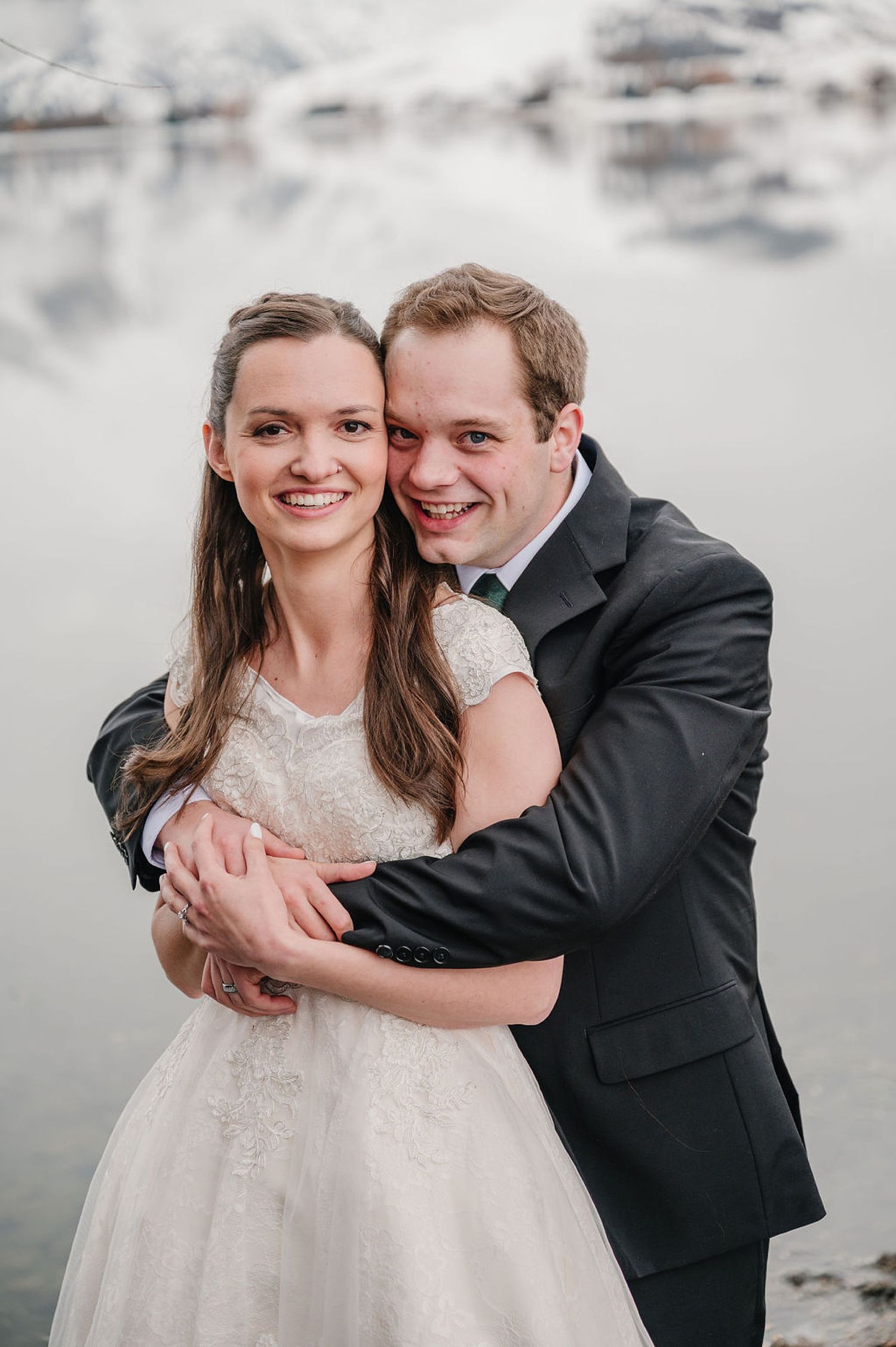 A romantic wedding portrait of a couple embracing by a waterfront in formal white dress and black suit.