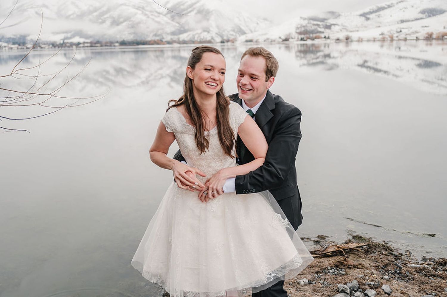 A couple shares a joyful moment in winter by a snowy mountain lake, the bride wearing a white tulle dress.