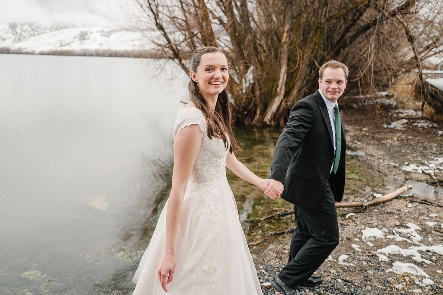 A couple in wedding attire stands by a snowy winter lake, with the bride in white and groom in dark suit.